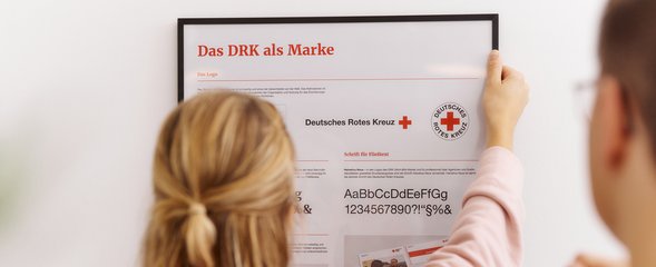 Abbildung zeigt Frau vor einem Plakat mit den Gestaltungselementen des DRK
